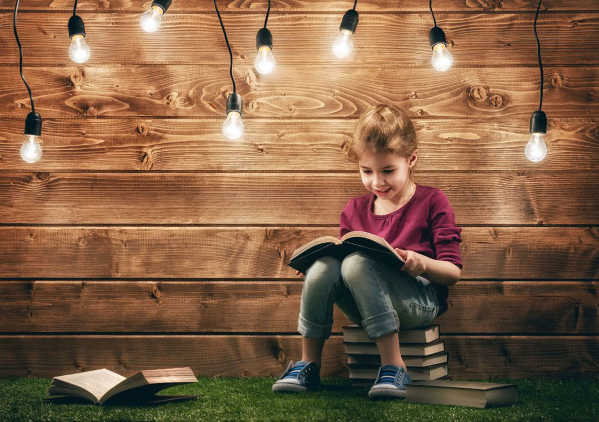 preschool girl reading book while sitting on pile of books